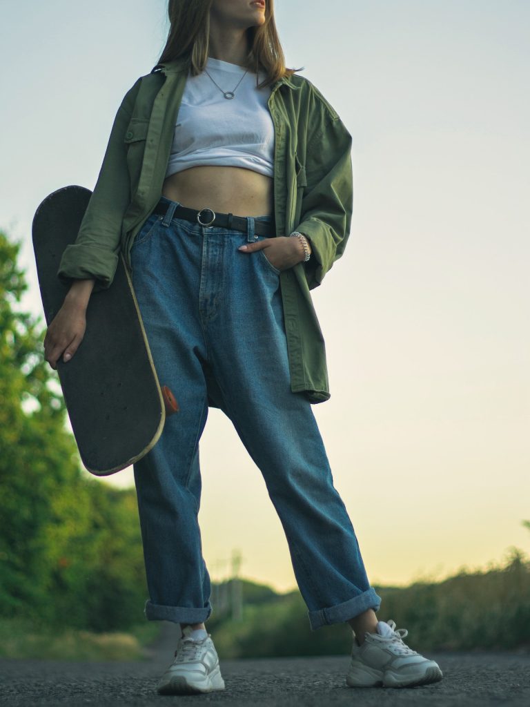 woman holding skateboard wearing white top green jacket and blue jeans
